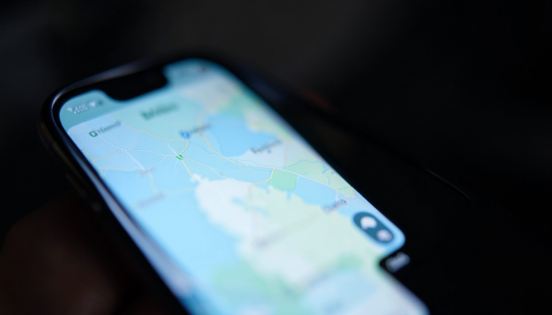 A close-up of a smartphone screen displaying a map interface with soft blue and green tones, set against a dark blurred background